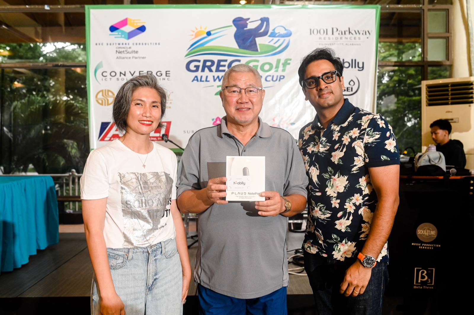 A raffle prize presentation in front of the GREC Golf All Weather Cup 2026 sponsor backdrop