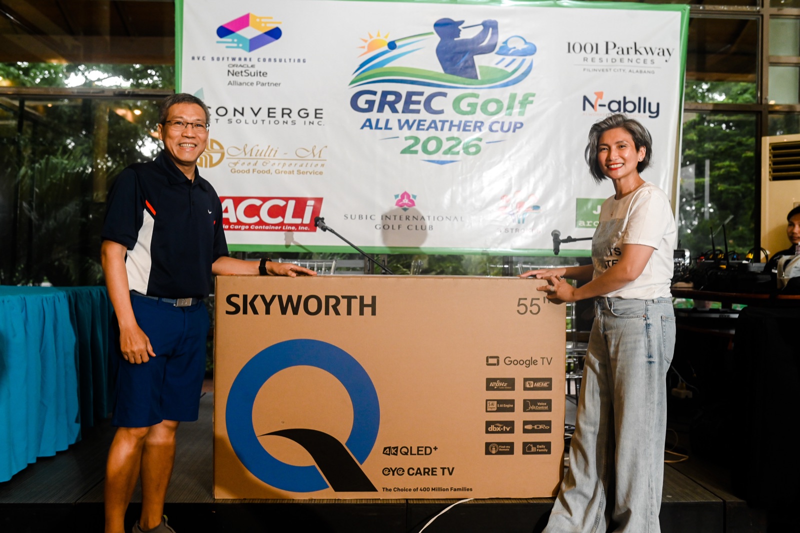 A raffle prize presentation in front of the GREC Golf All Weather Cup 2026 sponsor backdrop